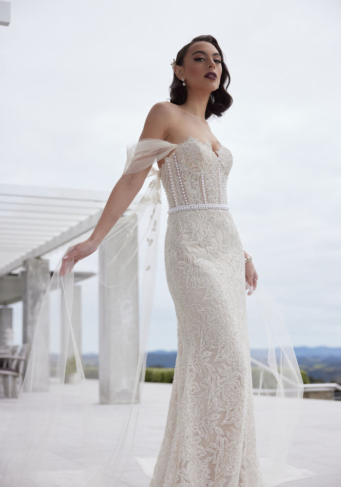 Tristan gown in champagne beading with pearl columns and waistband. Decorative tulle sleeves that flow into wings. Sweetheart strapless bodice and sheath silhouette.