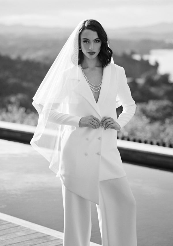 Black and white image of Ivory wedding blazer in asymmetric double-breasted cut. Worn with ivory trousers and veil.