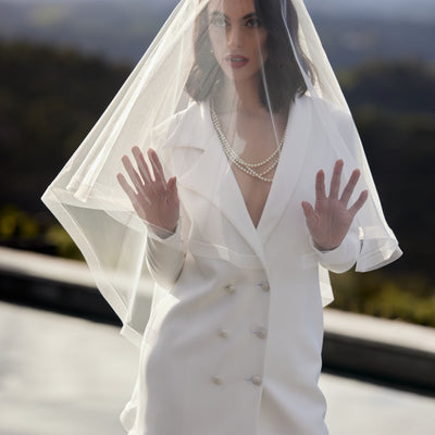 Ivory wedding blazer in asymmetric double-breasted cut. Worn with ivory trousers and veil over the face.