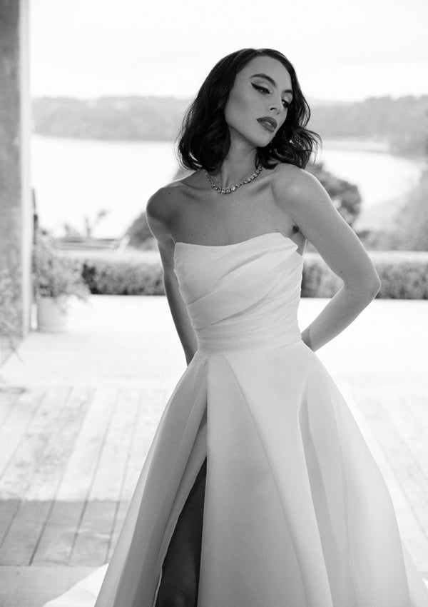 Black and white image of Tiffany gown. Ruched strapless bodice with full a-line skirt that opens into dramatic split from pleating detail.