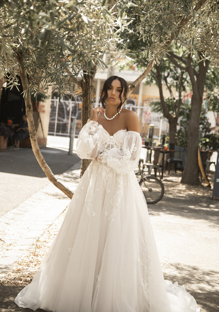 Soft tulle Rhoda dress in a-line silhouette with long tulle train. Bustier with visible boning and 3D lace applique. Detached balloon sleeves which billow from upper arm to a wide cuff at hem.