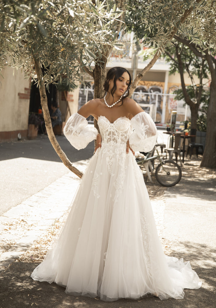 Soft tulle Rhoda dress in a-line silhouette with long tulle train. Bustier with visible boning and 3D lace applique. Detached balloon sleeves which billow from upper arm to a wide cuff at hem.