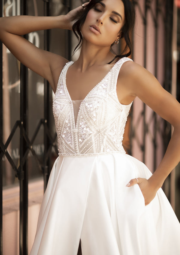 Ivory Raven gown with plunging v-neckline and dramatic a-line skirt with split. Bodice features geometric bead work and small mesh panel over bust for modesty.