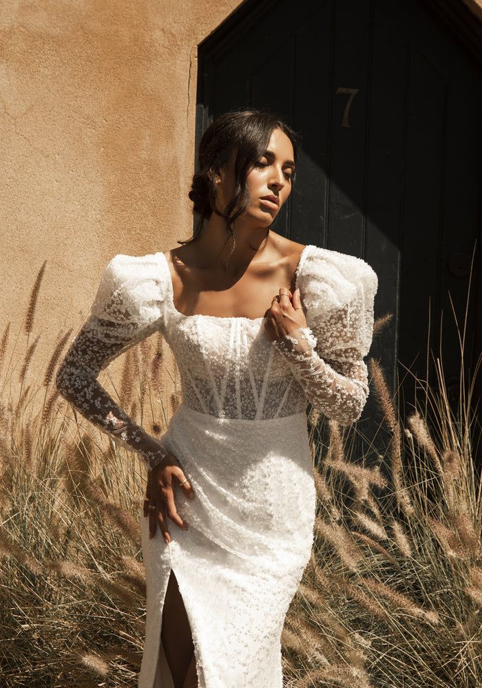 Fully beaded Rachel gown with sheer mutton sleeves and visible boning through the bodice. The ruched skirt is lined in ivory with asplit over the right leg.