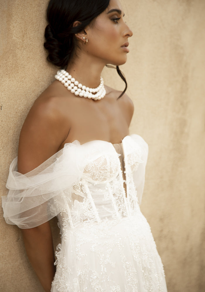 Close up of Rohini bodice with ruched tulle paneling, textured applique and ivory boning. Nude illusion mesh creates a plunging sweetheart neckline with off-the-shoulder tulle sleeves.